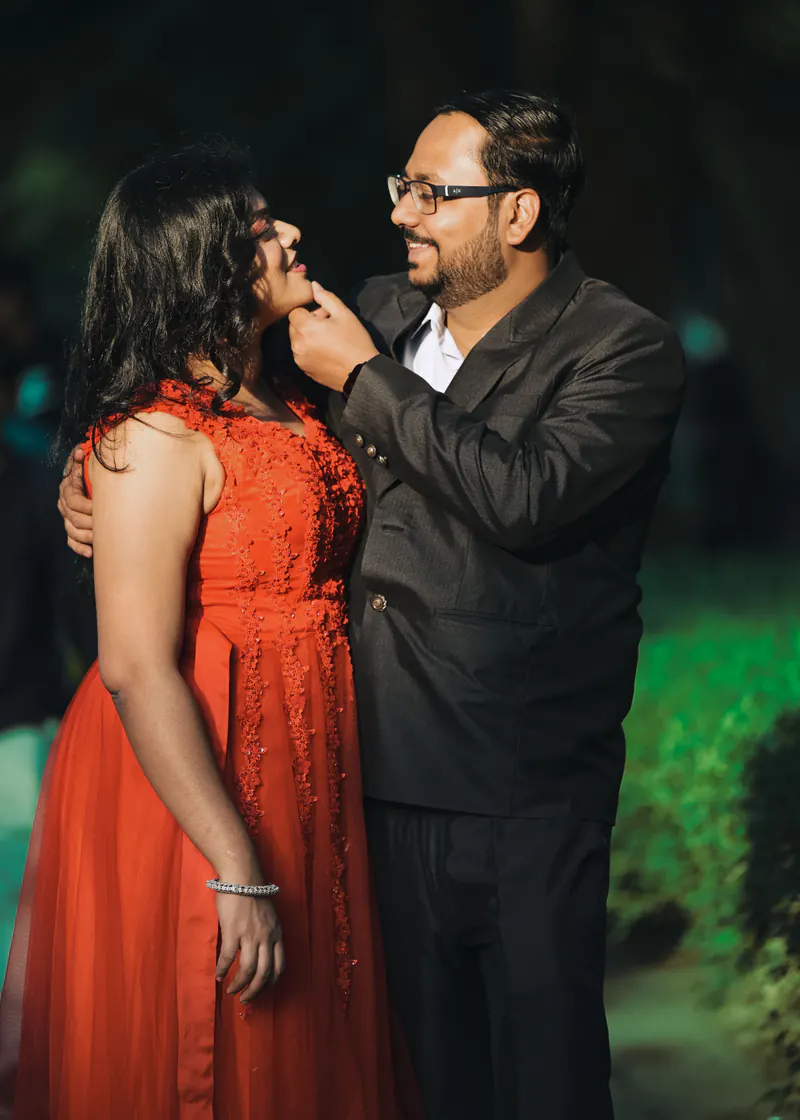 Couple at night, she in an orange dress lifts a hand to his face as he smiles in a dark jacket and glasses.