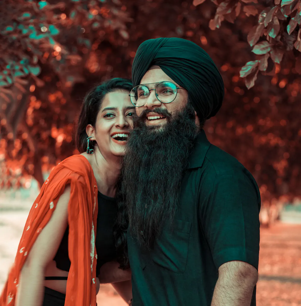 Sikh groom in a turban and glasses laughs beside his partner in an orange dupatta, against red autumn leaves.