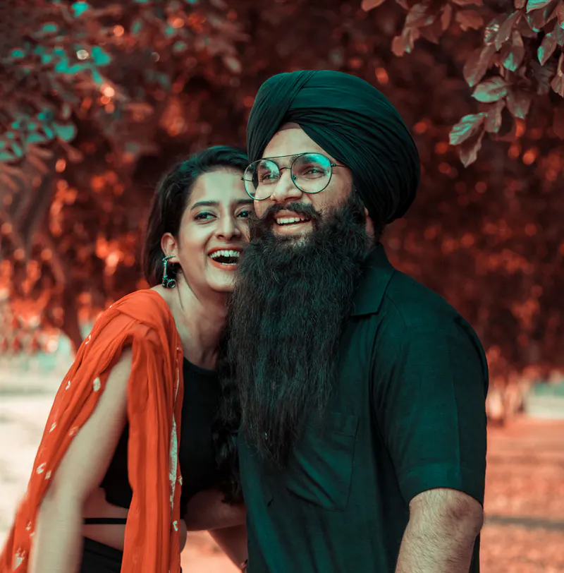 Sikh groom in a turban and glasses laughs beside his partner in an orange dupatta, against red autumn leaves.