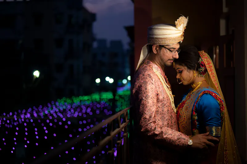 Groom in a safa and bride in a blue blouse and orange dupatta stand close on a balcony at dusk, purple lights below.