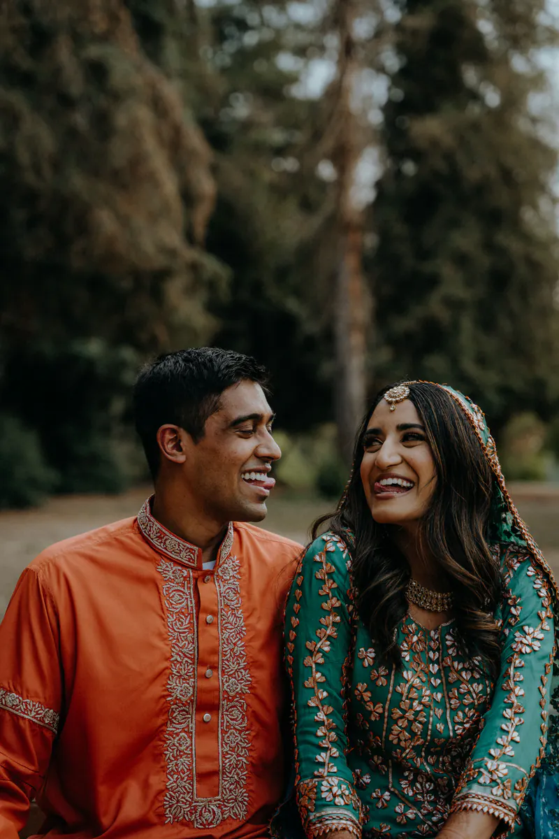 Couple seated outdoors among tall trees, he in an orange kurta and she in a teal lehenga, laughing together.