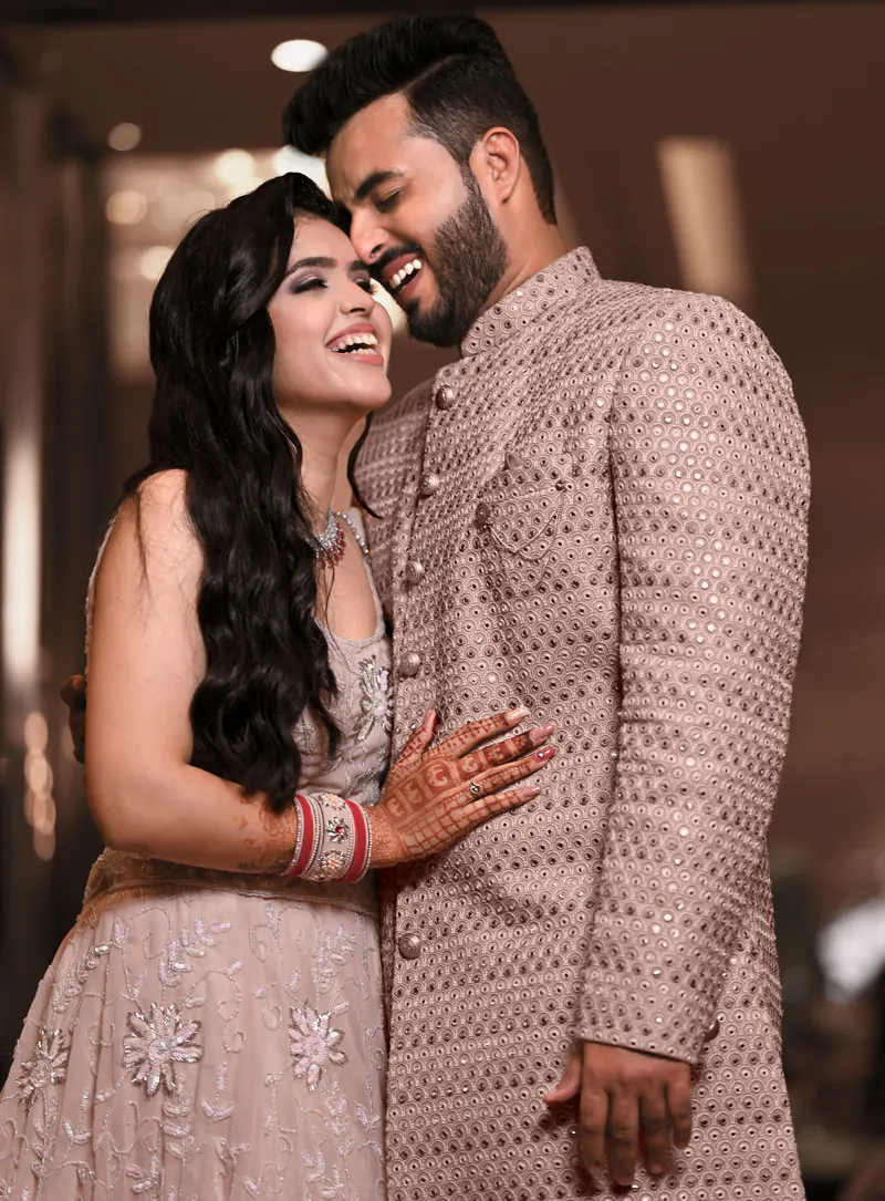 Bride in a blush lehenga and groom in a rose sherwani laugh with their heads together against warm indoor bokeh.