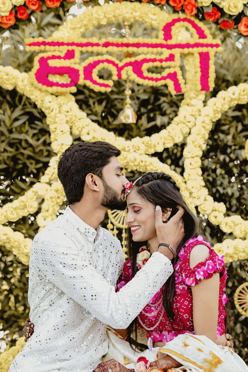 Groom kisses the bride on the forehead in front of a marigold haldi backdrop.