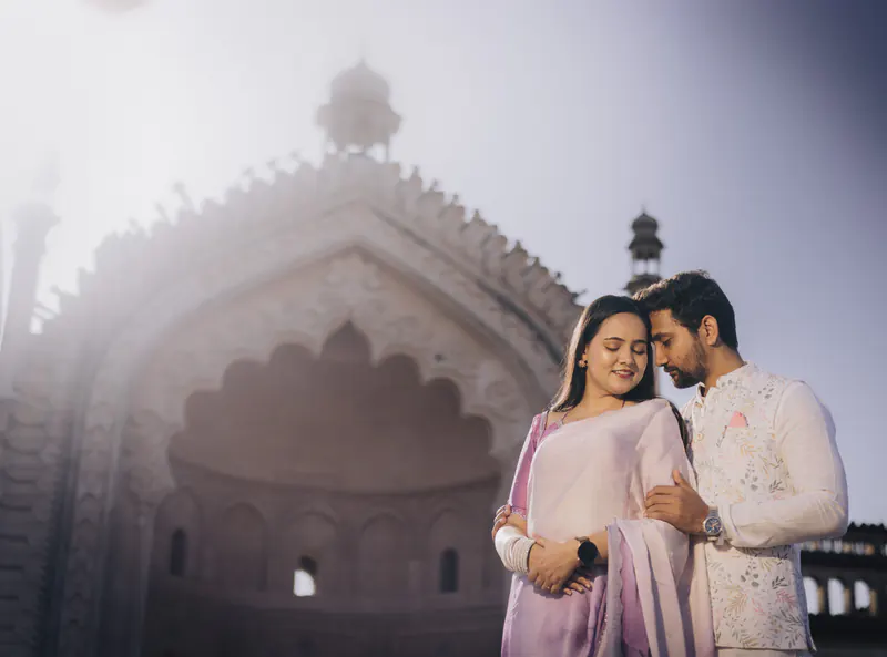 Couple stand close before a hazy Mughal monument arch, she in lilac, he in an ivory kurta.