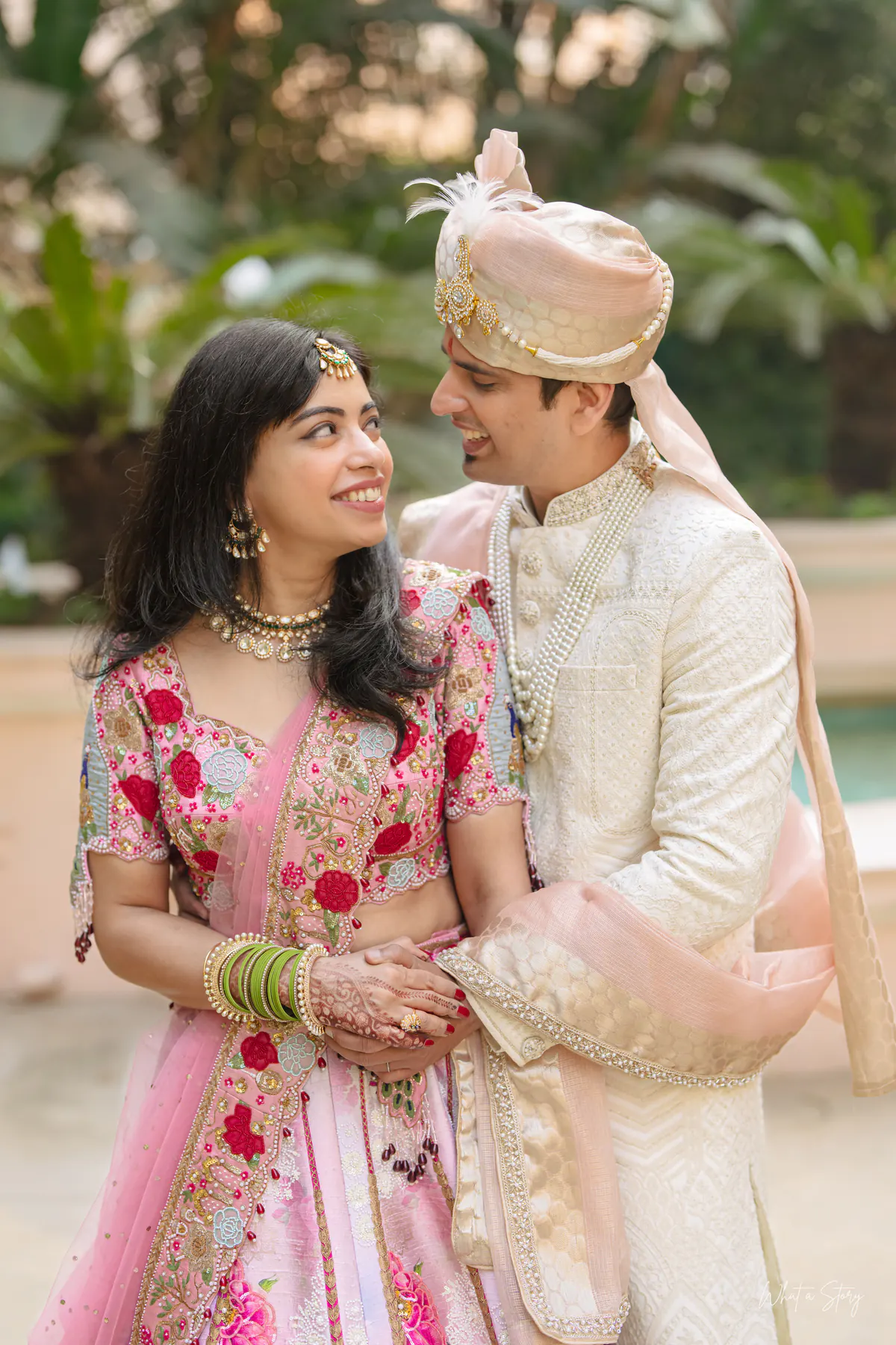 Bride in a pink floral lehenga and groom in a peach safa smile close together in a garden.