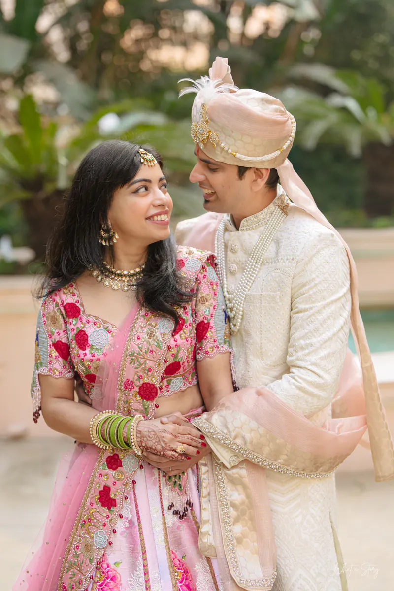 Bride in a pink floral lehenga and groom in a peach safa smile close together in a garden.