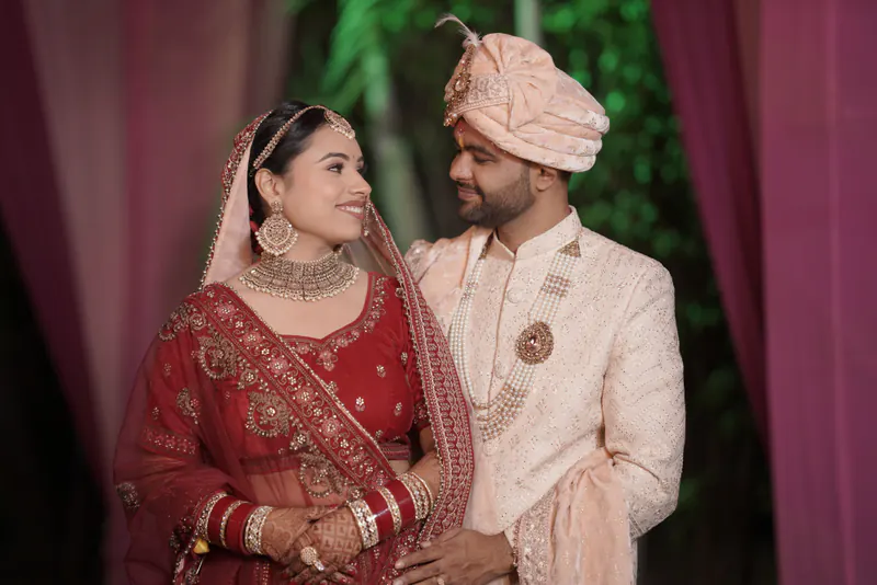 Bride in a red lehenga and groom in a pink safa and ivory sherwani smile in front of maroon drapes.