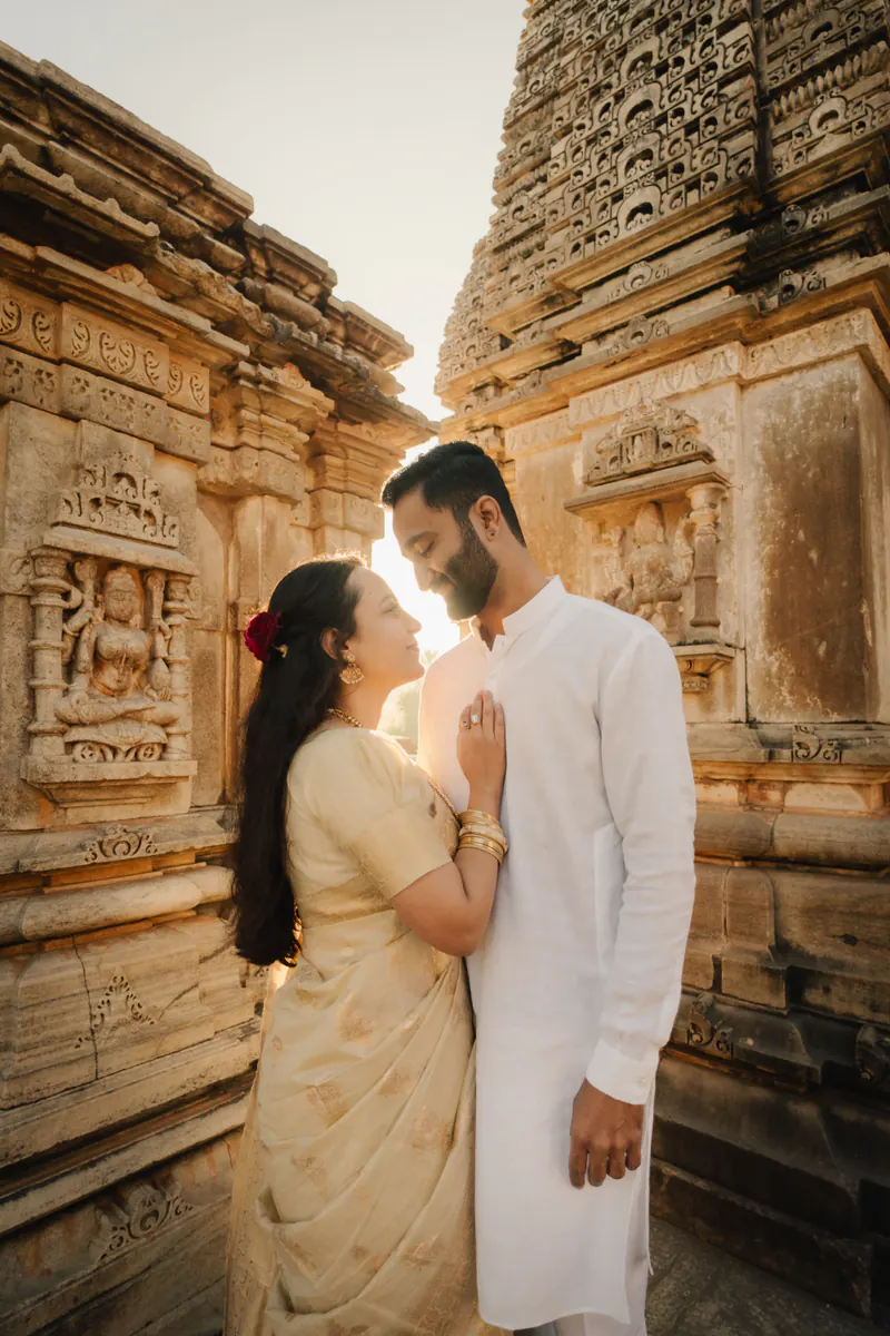 Couple face each other in a sunlit sandstone temple, she in a cream saree, he in a white kurta.