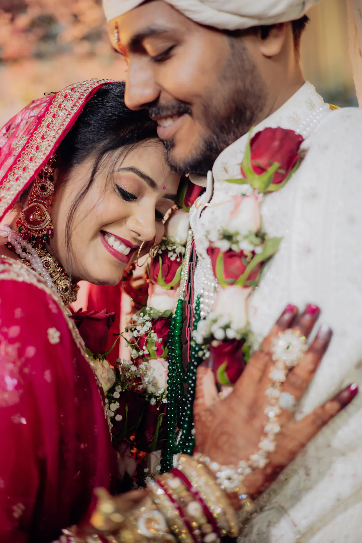 Close crop of a smiling bride with eyes closed and the groom resting his face against her head, a red rose in front.