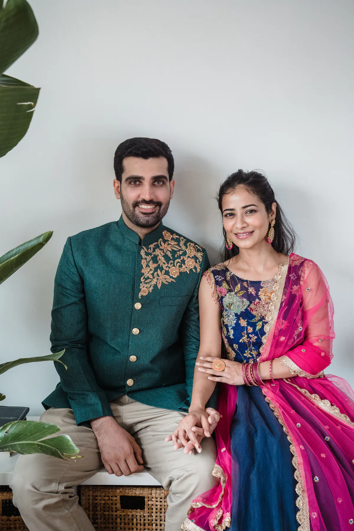Couple seated against a white wall with plants, he in a green sherwani, she in a blue and pink lehenga.