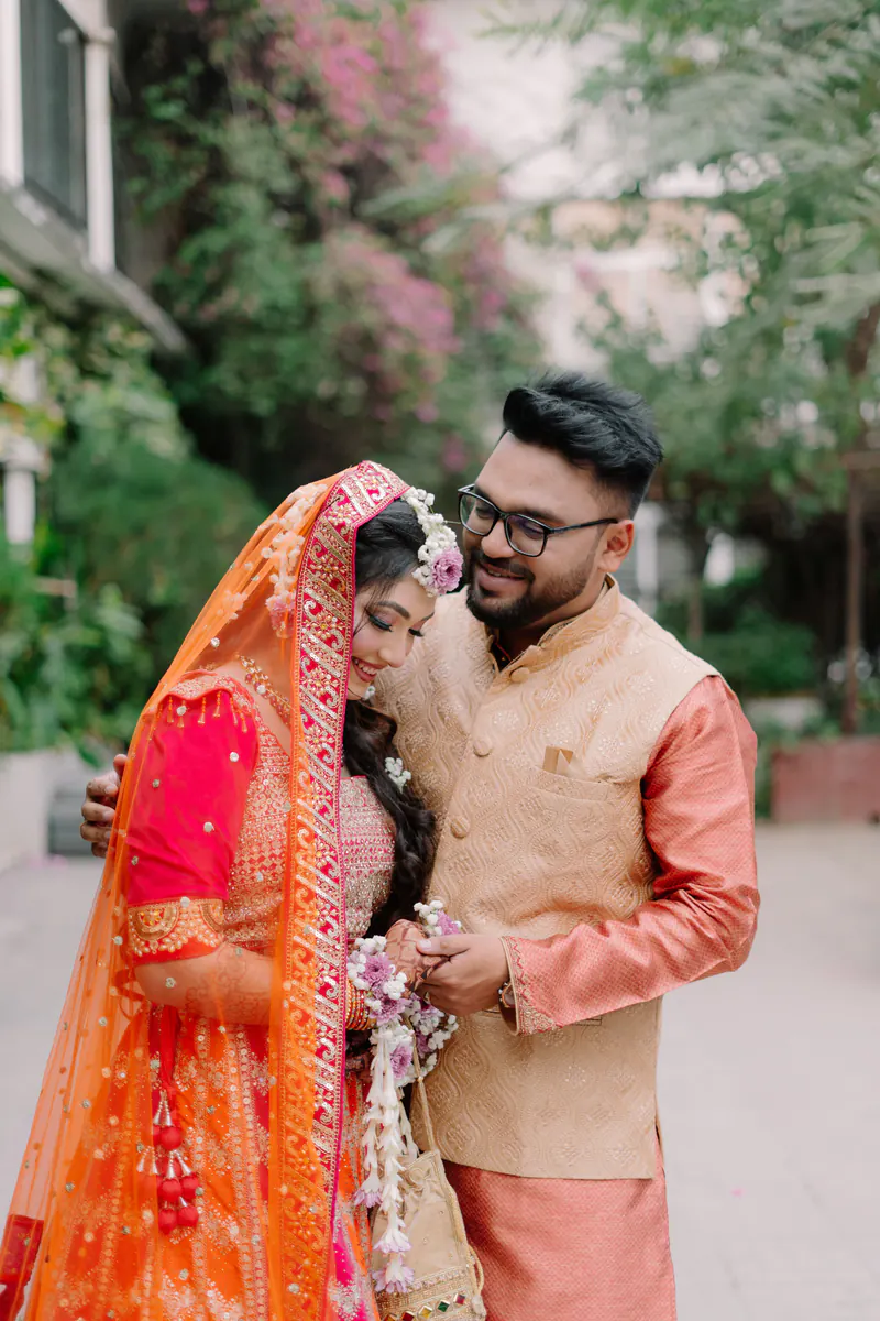 Bride in an orange and pink lehenga leans on the groom in a peach jacket and glasses in a leafy garden.