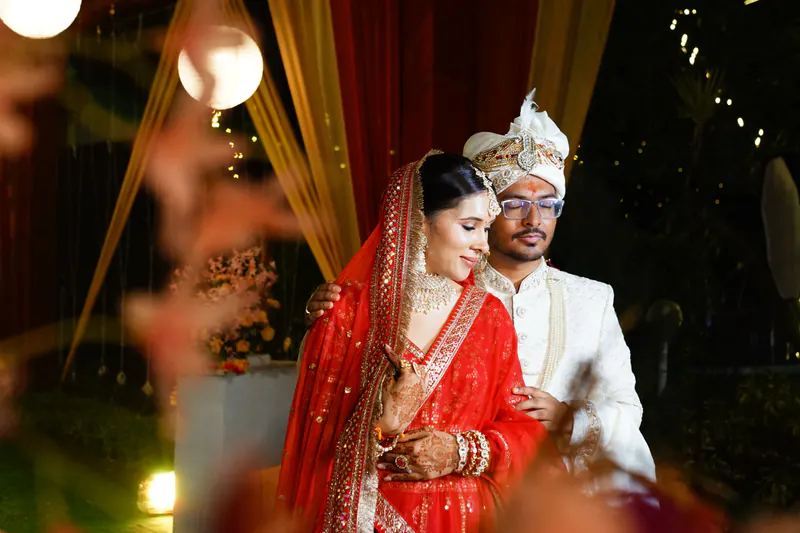 Bride in a red saree and groom in a white safa stand close at night, warm bokeh lights and drapes behind.